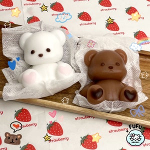 May include: Two plush toy bears, one white and one brown, are sitting on white gauze pads. The bears have black eyes and pink noses. The bears are surrounded by a white background with a strawberry pattern.