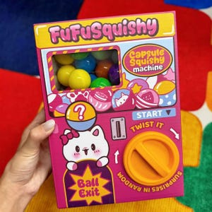 May include: A pink and yellow FufuSquishy capsule toy machine with a clear window showing colorful balls. The machine features a cute cat graphic, the text "Ball Exit", and instructions to "Twist It".