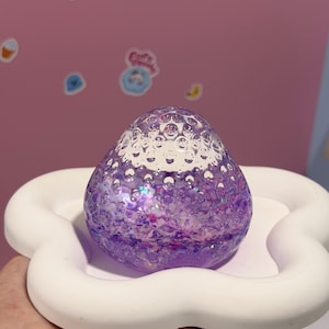 May include: A decorative, purple, strawberry-shaped object with a clear, bubble-textured top. The object is filled with glitter and sits in a white, cloud-shaped dish. The background is a light pink color.