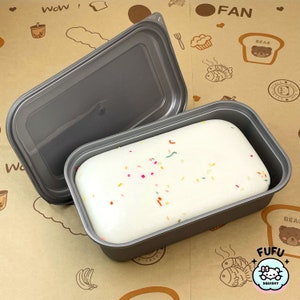 May include: A white cake-like slime with colorful sprinkles in a gray plastic container with a lid. The container is open and the slime is visible.