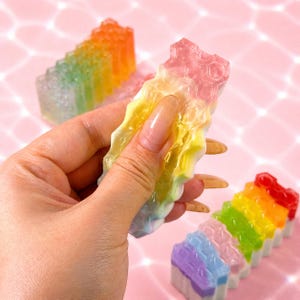 May include: A translucent, multi-colored, rectangular bar with a textured, honeycomb-like surface pattern. The bar features a gradient of colors including pink, yellow, green, and blue, and is held in a vertical position, showing a semi-flexible texture.