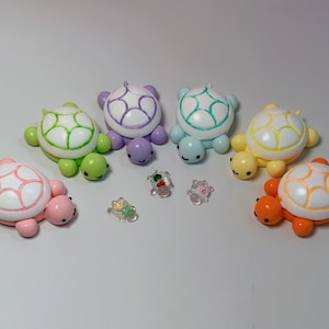 May include: A collection of colorful turtle figurines in various shades of pink, green, purple, blue, and yellow. Each turtle has a white shell with colored accents. Small, clear, decorative turtle-shaped beads are also present.