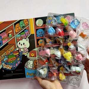 May include: A colorful illustration of a cartoon cat pushing a shopping cart filled with groceries. Next to it, a clear plastic tray holds miniature sushi and other food-themed figurines. The illustration features the text "Juice", "Chips", and "Fufu".