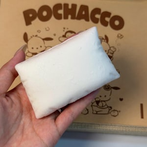 May include: A small, rectangular, plain white pillow-shaped object held in front of a tan cardboard box. The box features the brown text "POCHACCO" at the top, accompanied by brown line-art illustrations of the cartoon character Pochacco.