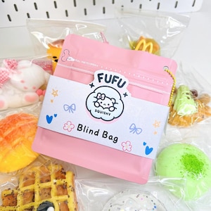 May include: A pink zippered pouch with the text "Fufu Squishy Blind Bag" and a cartoon cat illustration. The pouch is surrounded by various colorful squishy toys, including a waffle, a peach, a green and white swirl, and a white bunny.