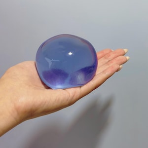 May include: A translucent, blue-purple sphere, held in an open hand. The sphere has a smooth, gel-like appearance, with subtle color variations. The background is a plain, light gray.