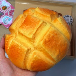 May include: A round, golden-brown squishy toy shaped like a loaf of bread with a textured, segmented top surface. The object features a realistic baked appearance with deep grooves and a soft, porous-looking finish, mimicking a freshly baked bun.