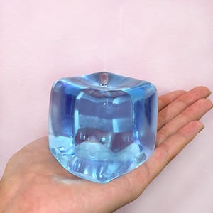 May include: A clear, blue, cube-shaped decorative object with rounded edges. The object has a small, rounded top and is held in a hand. The object is made of a transparent material, reflecting light and creating a shimmering effect. The background is a soft pink.