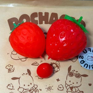 May include: Two large red strawberry-shaped squeeze toys with green plastic stems and one small red spherical toy. The items are positioned on a light brown paper surface featuring brown line art illustrations of cartoon characters.