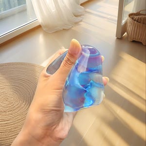 May include: A clear, iridescent glass object with blue and purple hues. The object is held in a hand, showcasing its smooth, organic shape. The glass reflects light, creating a shimmering effect. The background includes a window and a woven basket.