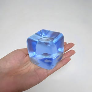 May include: A translucent blue square-shaped object with a textured surface. The object is held in a hand against a white background. The object appears to be made of glass or a similar material.