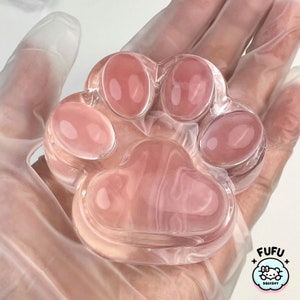 May include: A clear pink paw-shaped squishy toy with a textured surface. The toy is made by Fufu Squishy.