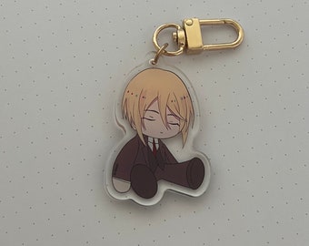 May include: A clear acrylic keychain featuring a cartoon character with blonde hair, wearing a dark suit and sitting with their legs crossed. The keychain has a gold metal ring.