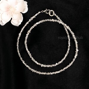 May include: A delicate necklace composed of small, irregularly shaped, light gray gemstones. The necklace is double-stranded and secured with a silver-colored clasp. The background is black, with a white flower in the upper left corner.
