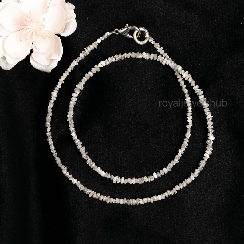 May include: A delicate necklace composed of small, irregularly shaped, light gray gemstones. The necklace is double-stranded and secured with a silver-colored clasp. The background is black, with a white flower in the upper left corner.