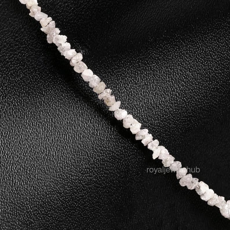 May include: A strand of rough-cut, raw diamond beads. The diamonds are a light, off-white color and appear to be irregularly shaped. The beads are strung together, creating a necklace or bracelet. The background is a dark, textured surface.