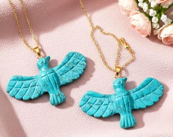 May include: Two turquoise bird pendant necklaces with gold chains. The pendants are carved with detailed wing and feather patterns. The necklaces are displayed on a pink fabric surface, with a bouquet of pink roses in the background.