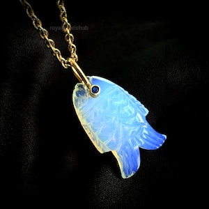 May include: A gold-toned chain necklace with a pendant shaped like a fish. The fish pendant is carved from a light blue and white material, with a small blue gem set as an eye. The background is black.