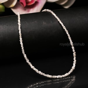 May include: A delicate necklace composed of small, irregularly shaped, light-colored gemstones. The necklace is displayed against a black background, with a hint of a pink flower in the upper left corner. The gemstones appear to be raw diamonds.