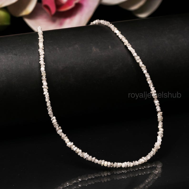 May include: A delicate necklace composed of small, irregularly shaped, light-colored gemstones. The necklace is displayed against a black background, with a hint of a pink flower in the upper left corner. The gemstones appear to be raw diamonds.