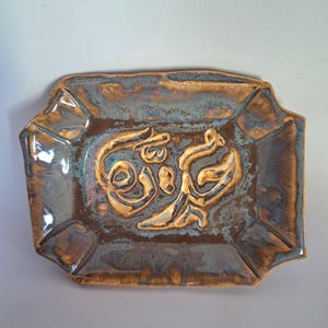 May include: A rectangular ceramic tray with a raised, ornate design in gold against a mottled brown and blue background. The tray has a slightly irregular shape with angled edges.