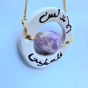 Könnte beinhalten: Eine Halskette mit einem weißen Keramikring-Anhänger, der mit schwarzer arabischer Kalligrafie beschriftet ist. Ein glatter, polierter, hellvioletter Amethyststein ist in der Mitte des Rings mit goldfarbenen Metallringen an einer Goldkette befestigt.