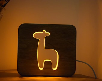 Op de afbeelding: Houten nachtlampje met een girafsilhouet. De girafvorm wordt verlicht met een warme, zachte gloed. De lamp is vierkant en heeft een natuurlijke houtnerf afwerking. Een witte kabel loopt van de achterkant.