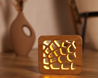 May include: A square wooden lamp with a geometric cutout design. The lamp emits a warm, yellow glow, creating a cozy ambiance. A brown vase with dried plants is in the background, suggesting a home decor theme.