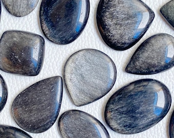 Könnte beinhalten: Eine Sammlung polierter Obsidian-Edelsteine in verschiedenen Formen, darunter oval, tropfenförmig und quadratisch. Die Steine haben eine dunkle, reflektierende Oberfläche mit silberfarbenen Einschlüssen, die einen schimmernden Effekt erzeugen. Die Edelsteine sind auf einer weißen Oberfläche angeordnet.