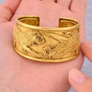 May include: A gold-colored metal cuff bracelet featuring a raised, textured relief design of two tigers facing each other. The bracelet has an open back design and a polished finish, showcasing detailed fur patterns and facial features on both animals.