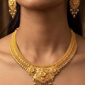Puede incluir: Un conjunto de collar y pendientes colgantes de oro con intrincados motivos florales, metal texturizado y delicadas cuentas esféricas colgantes. El collar tiene una banda ancha y estructurada con un diseño floral central y flecos.