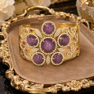 May include: A gold cuff bracelet featuring five large, round, purple cabochon stones arranged in a floral pattern. The bracelet is accented with small, clear pavé-set stones, intricate white scrollwork, and small, round, pink accent stones.