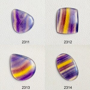 Könnte beinhalten: Vier Fluoritsteine in verschiedenen Formen und Größen. Die Cabochons sind lila, gelb und weiß mit einem Bandmuster. Die Cabochons sind mit 2311, 2312, 2313 und 2314 beschriftet.