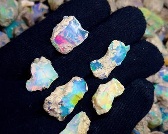 Könnte beinhalten: Eine Sammlung roher Opale mit leuchtenden Farben wie Blau, Grün, Rosa und Gelb. Die Opale werden in einem schwarzen Handschuh gehalten, mit dem Text "STONERIAGEMS" am unteren Rand.