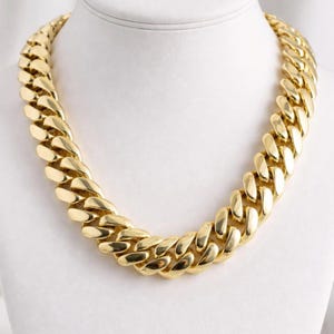 Puede incluir: Un collar de cadena grueso en tono dorado pulido con eslabones de bordillo redondeados y entrelazados. El collar tiene un acabado metálico altamente reflectante y brillante, con una apariencia robusta y pesada en forma circular.