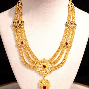 Puede incluir: Un collar de oro de varias hebras con eslabones de filigrana intrincados, acentuado con piedras preciosas rojas redondas y pequeños detalles de piedras verdes. El colgante central es un elemento de oro en forma de flor con una piedra roja.