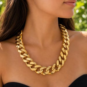 Puede incluir: Un collar de cadena grueso en tono dorado con eslabones grandes e interconectados. El collar se ajusta al cuello, mostrando una superficie metálica reflectante con un color amarillo dorado brillante y uniforme en todo el diseño.