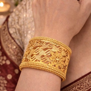 Puede incluir: Una pulsera brazalete ancha de oro con un intrincado diseño calado de líneas onduladas y un motivo floral prominente, bordeada por bordes de cuentas. Un anillo de banda de oro fino a juego se lleva en el dedo, complementando el trabajo.
