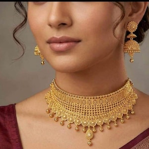 Puede incluir: Un collar tipo gargantilla de oro con un intrincado patrón de filigrana, textura de cuentas y dijes colgantes en forma de lágrima en el borde inferior, junto con pendientes jhumka a juego en forma de campana con un acabado dorado texturizado.