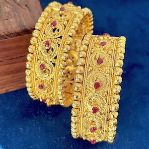 Puede incluir: Dos brazaletes de oro con intrincados patrones de filigrana y pequeñas piedras preciosas rojas en forma de lágrima. Los bordes de cada brazalete están delineados con un diseño texturizado de cuentas, creando una superficie metálica detallada.