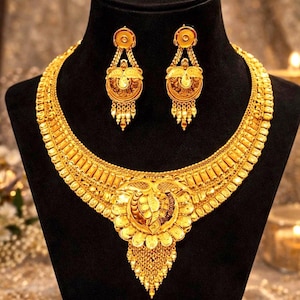 Puede incluir: Un collar de oro y un par de pendientes a juego. El collar presenta una banda ancha y texturizada con un gran colgante central adornado con grabados intrincados y flecos colgantes. Los pendientes reflejan el diseño del colgante central.