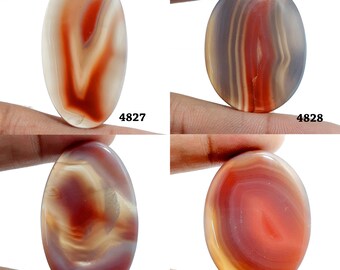 Könnte beinhalten: Vier ovale Achat-Cabochons mit unterschiedlichen Mustern in Rot, Weiß und Braun. Die Cabochons werden auf einer Hand gezeigt.