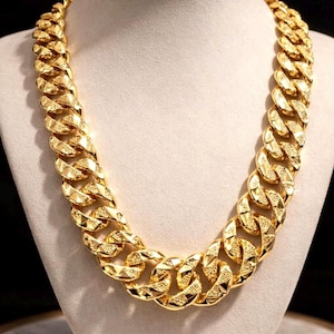 Puede incluir: Un collar de cadena gruesa de color dorado con eslabones grandes e interconectados. Cada eslabón tiene una superficie texturizada y facetada que refleja la luz, creando una apariencia metálica brillante en toda la pieza de joyería.