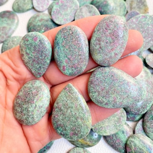Peut inclure: Une collection de cabochons de zoïsite rubis vert et rose, avec une main tenant plusieurs pierres. Les pierres sont lisses et polies, avec une variété de formes et de tailles.