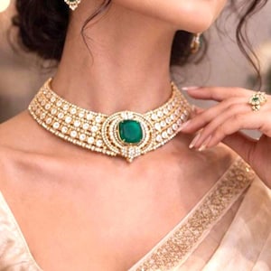 Puede incluir: Un collar tipo gargantilla en tono dorado con tres filas de pequeñas piedras transparentes y una gran piedra central de color verde esmeralda en talla cojín, rodeada por un marco decorativo de piedras más pequeñas, con un anillo a juego.