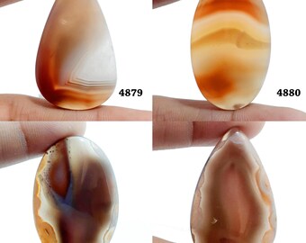 Könnte beinhalten: Vier Achat-Cabochons in verschiedenen Brauntönen, Orange und Weiß. Die Cabochons sind tropfenförmig und haben eine polierte Oberfläche. Die Cabochons sind mit den Nummern 4879, 4880, 4881 und 4882 gekennzeichnet.