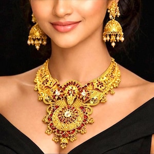 Puede incluir: Un conjunto de collar y pendientes jhumka en tono dorado. El collar presenta un diseño de filigrana intrincado con piedras rojas incrustadas y detalles blancos. Los pendientes en forma de campana cuelgan de postes dorados con cuentas.