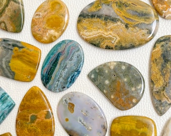 May include: A collection of polished, natural gemstone cabochons in various shades of yellow, brown, gray, and blue. The stones have unique patterns and textures, showcasing the beauty of natural materials.