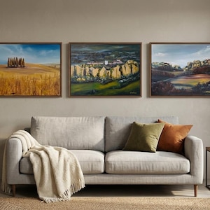 Può includere: Tre dipinti di paesaggi incorniciati sono appesi sopra un divano grigio con cuscini e una coperta. I dipinti raffigurano scene rurali con campi, alberi ed edifici. L'opera d'arte è esposta in un soggiorno.