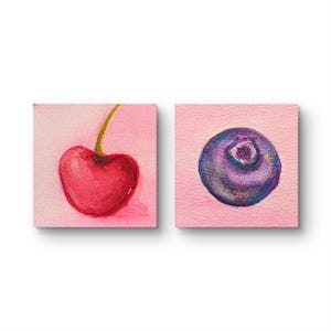 Könnte beinhalten: Aquarellmalereien einer Kirsche und einer Blaubeere auf quadratischen, rosa Hintergründen. Die Kirsche ist leuchtend rot mit einem grünen Stiel. Die Blaubeere ist tiefblau und lila mit detaillierter Textur.